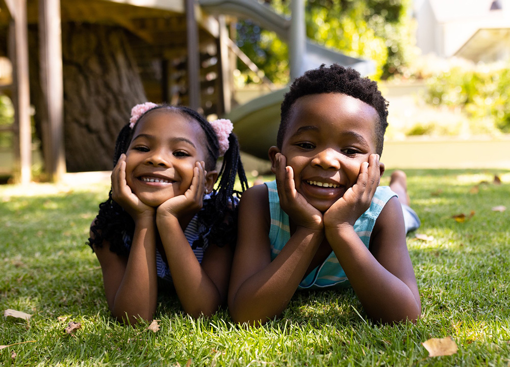 Two smiling schoolchildren laying on the grass in front of a playground